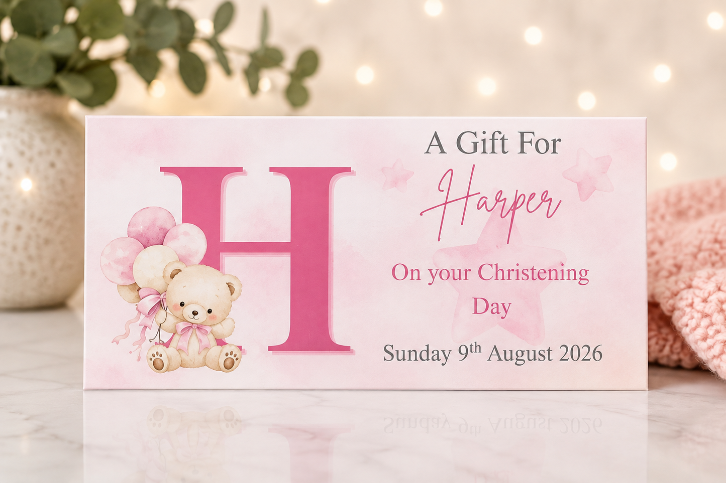 A personalized baby christening gift card featuring a large pink letter 'H', a cute teddy bear holding balloons, and the text 'A Gift For Harper On your Christening Day Sunday 9th August 2026'. The card is styled on a marble surface with a soft pink knit blanket and bokeh fairy lights in the background.