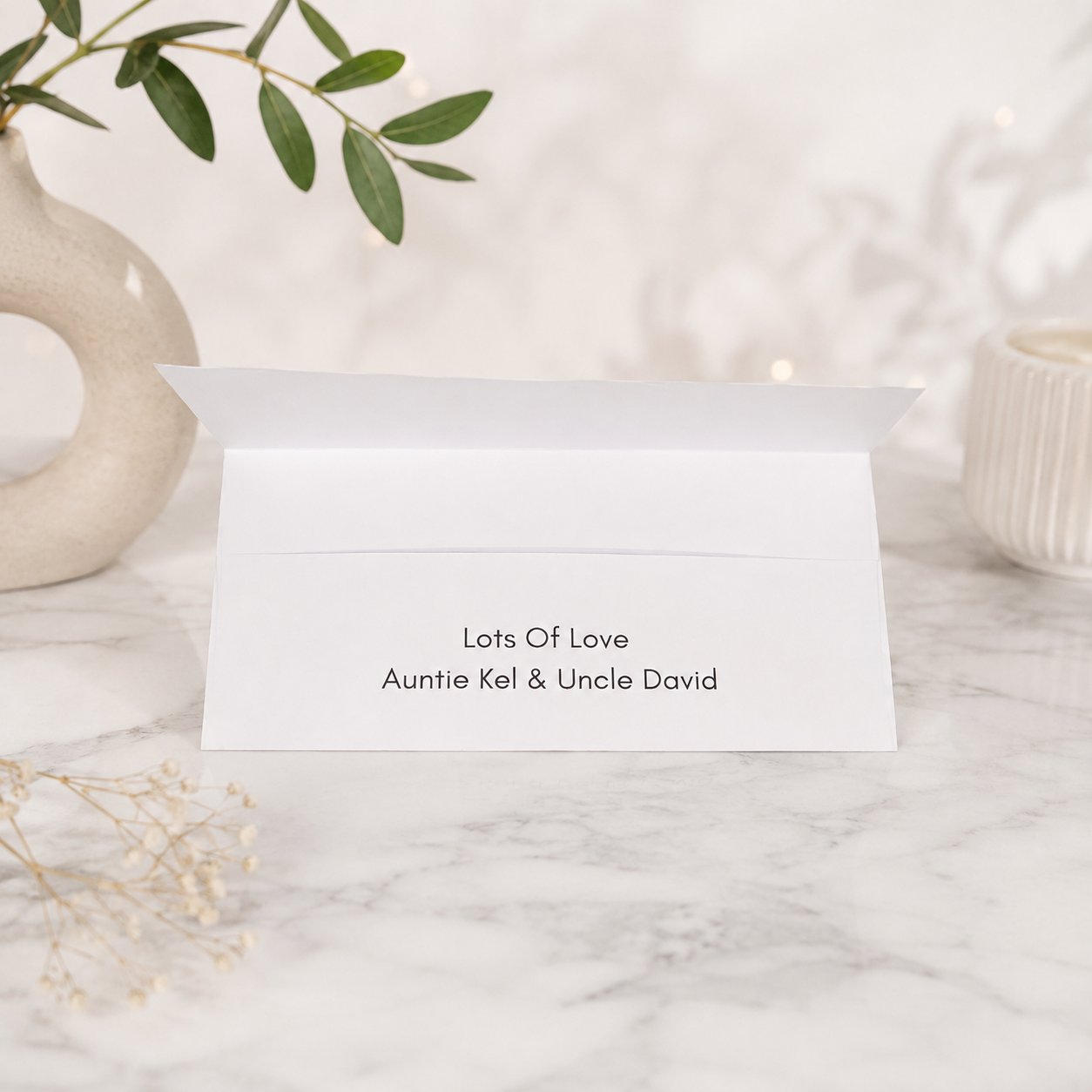 A white folded greeting card or envelope standing open on a marble surface, printed with the text 'Lots Of Love Auntie Kel & Uncle David.' Decorative ceramic vessels, greenery, and dried flowers are softly blurred in the background.