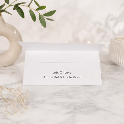 A white folded greeting card or envelope standing open on a marble surface, printed with the text 'Lots Of Love Auntie Kel & Uncle David.' Decorative ceramic vessels, greenery, and dried flowers are softly blurred in the background.